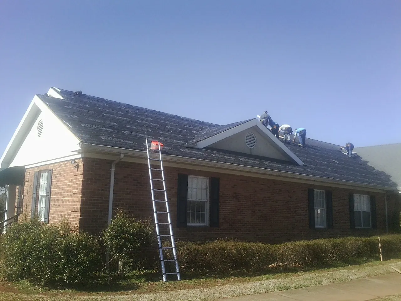 Crew tearing shingles off a brick ranch roof with a ladder set — Emergency Roof Repair work by our crew in Alum Bank