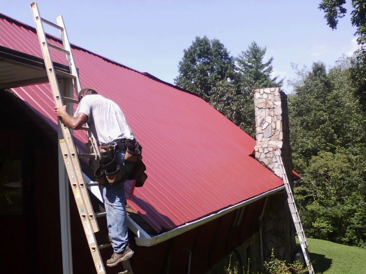 Roofer on a ladder beside a red metal roof and stone chimney — Soffit & Fascia Repair by Roofing Brothers in Alum Bank